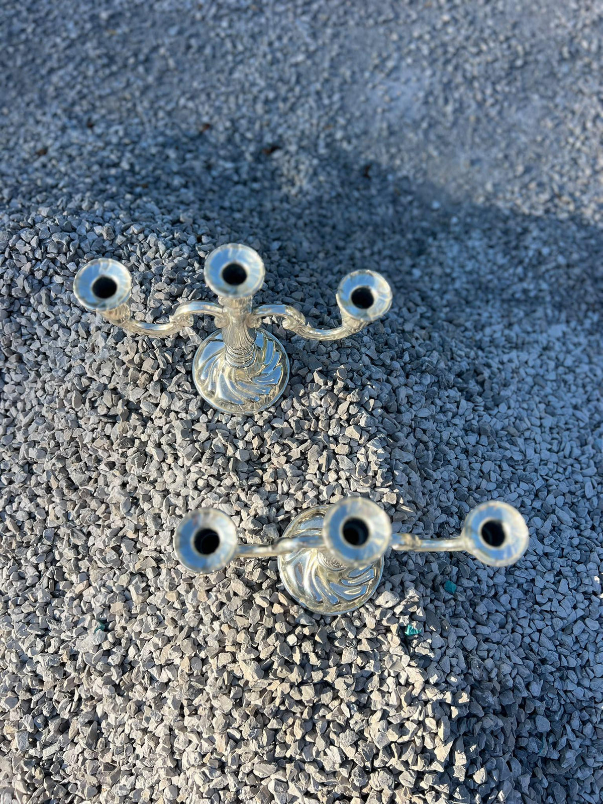 La Ferme Anglaise pair of ornate silver-tone metal 3-arm candelabra with scrolling designs on a gravel surface.